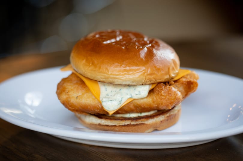 Golden-brown fried fish sandwich with cheese and tartar sauce on a brioche bun, on a white plate.