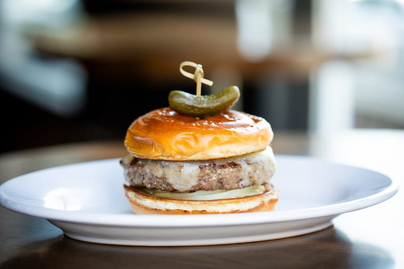 A gourmet cheeseburger with a pickle on top, served on a white plate.