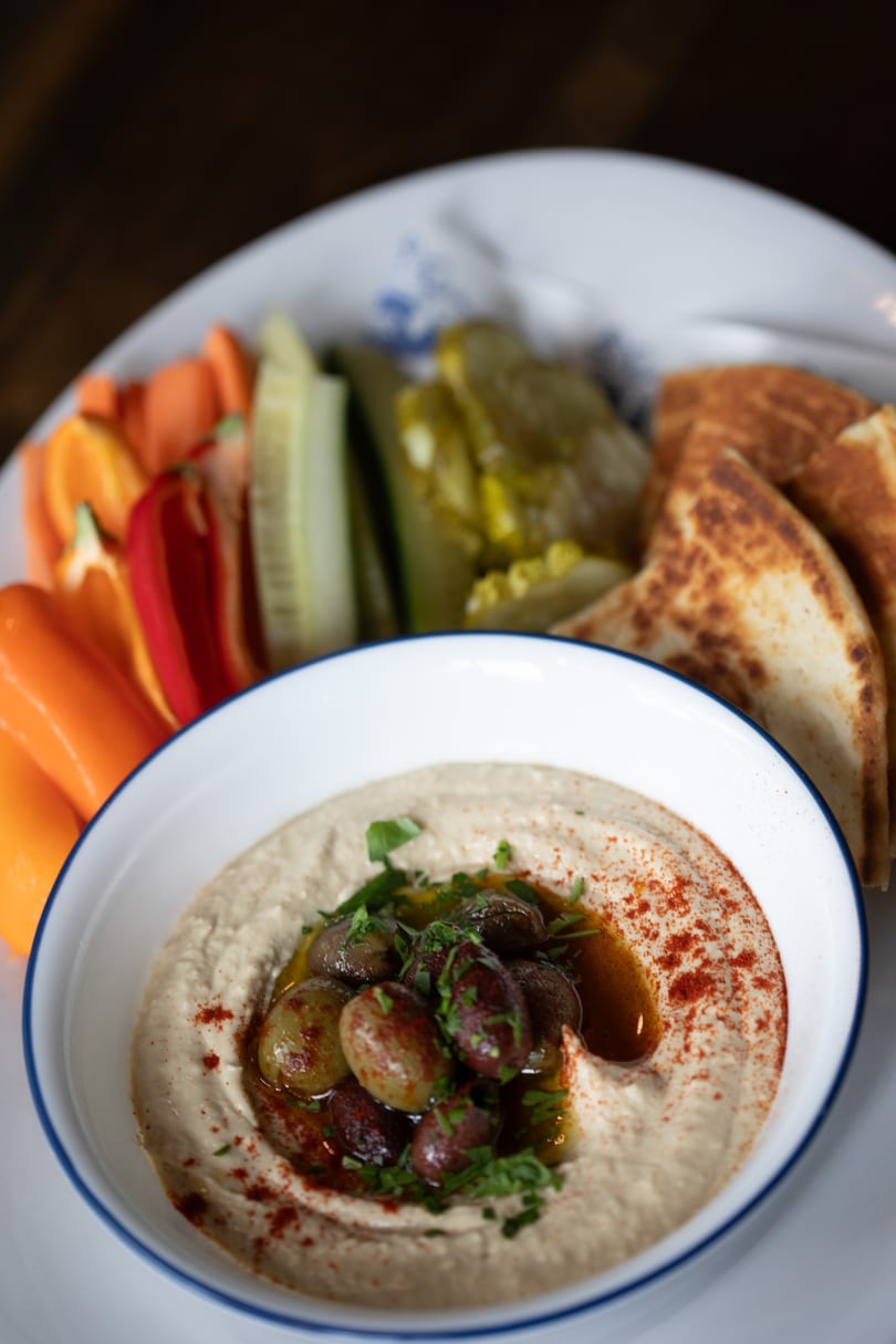 Hummus with olives, paprika, and parsley, served with pita bread and fresh vegetables.